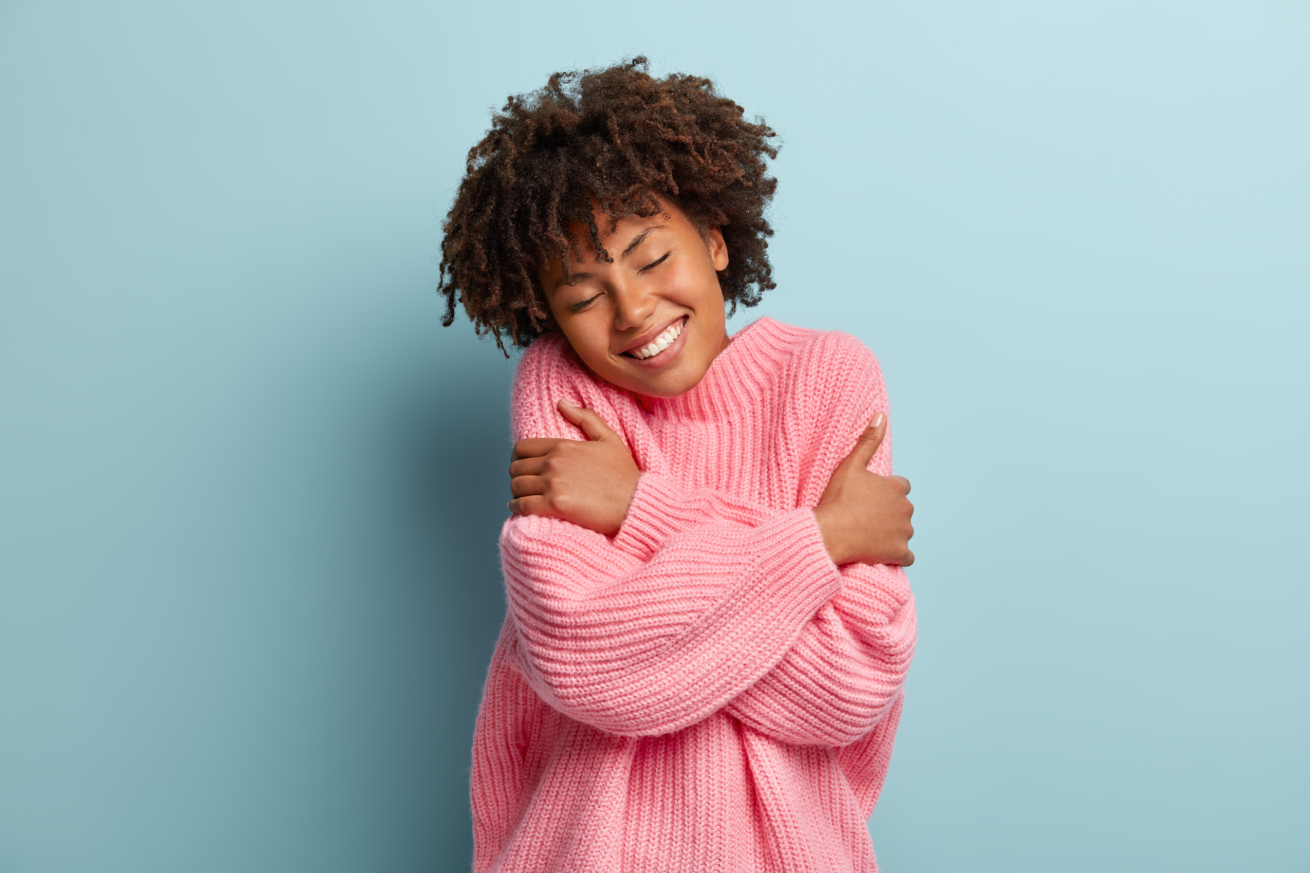 Love yourself concept. Photo of lovely smiling woman embraces herself, has high self esteem, closes eyes from enjoyment, likes her new comfortable soft pink sweater, tilts head, stands indoor